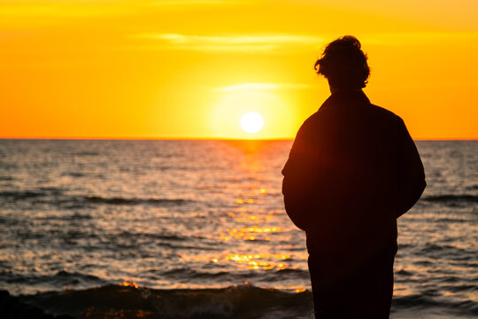 Dark silhouette of a man standing by the sea at golden sunset. Lonely teenager looking a setting sun.