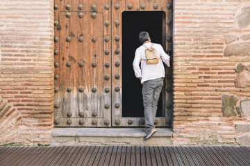 A middle aged man with a backpack looks out through an old open traditional wooden door. Rural and...