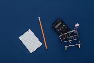 Shopping list. Shopping trolley with calculator, notebook and pencil on classic blue background. Color 2020. Top view