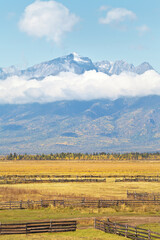 Beautiful calm rural autumn landscape with yellowed fields against the background of mountain snow-capped peaks with low clouds on a sunny day. Buryatia, Eastern Sayans, Baikal region, Tunka Valley