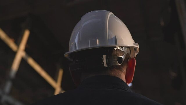 An engineer, builder, architect, man, holds a white protective helmet in his hands, looks into distance and puts it on his head. The manager at the facility watches the progress of the work performed