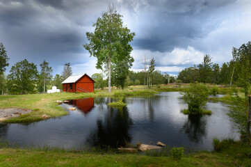 Obraz premium Small red Swedish house reflected in a small lake under a dramatic sky.