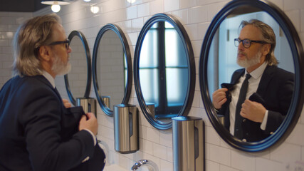 Elegant senior entrepreneur doing hair looking into mirror in office restroom