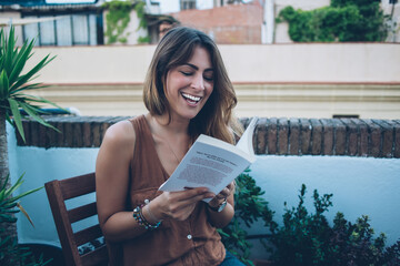 Cheerful adult lady reading funny book on balcony