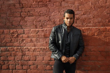 Young handsome stylish guy in a black leather jacket is leaning against the red brick wall outside.