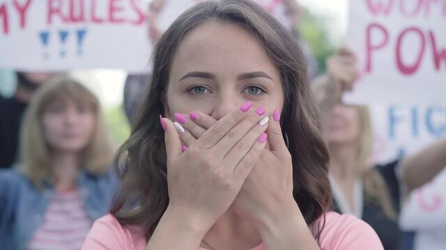 Close-up portrait of young beautiful woman closing mouth with hands as crowd of people protesting at the background. Female Caucasian activist showing no freedom of speech gesture on demonstration.