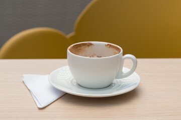 small white cup with chocolate, a plate and napkin, on a flat wooden surface, with a yellow chair in the background