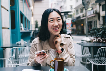 Laughing young woman browsing cellphone while enjoying coffee sitting in cafe on street