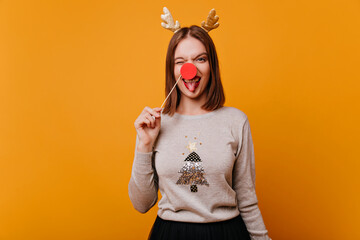 A bright, emotional girl with a Christmas mood in a fun outfit and accessories in the form of golden horns and a red deer nose looks into the camera and blinks.