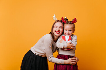 A smiling mother with red lips hugs a happy daughter holding a Christmas present in her hands. Photo of a young woman and child in hoops on the head with deer horns.