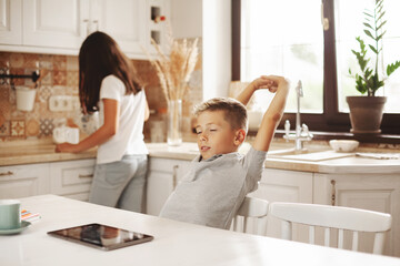 A tired boy pulls himself up after online lessons in the kitchen at home, his sister helps him. Distance learning, online education, home schooling concept