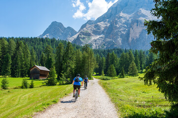 Group of mountain bikers riding their bicycles on a trail going through green meadows in the Italian Alps. Dolomite peaks are visible in the background. Val Badia, South Tyrol - Italy