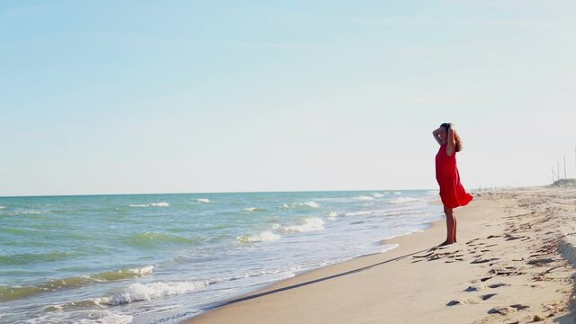 A beautiful girl in a red dress walks along the seashore. A young woman in a red dress walks along the beach.
