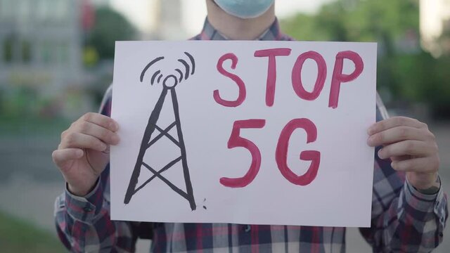 Camera approaches to Stop 5G placard in hands of Caucasian man in face mask. Male activist protesting against radioactive pollution of mobile towers causing Covid-19 respiratory disease.