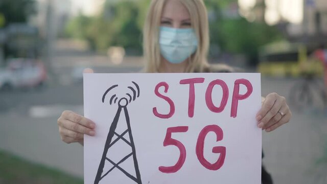 Blond Caucasian woman in face mask protesting against 5G mobile towers. Rack focus changes from female activist to banner with slogan Stop 5G. Individual riot against radiation causing Covid-19.