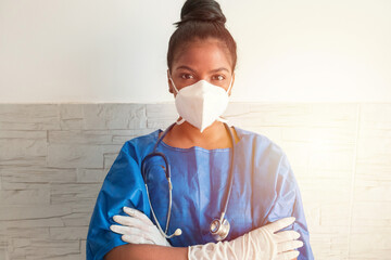 A young African nurse wears a blue uniform, a mask and gloves while standing with her arms crossed...