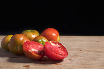 The tomato is cut in half. Tomatoes of different ripeness lie on a white oak board against a dark background. Vegetarian menu element.