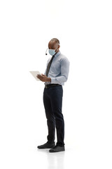 Works with gadgets in face mask. Young african-american call center consultant with headset on white studio background. Copyspace for ad, text. Concept of professional occupation, work, job
