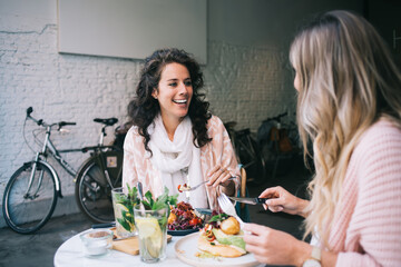 Cheerful caucasian female colleagues sitting together in cafe interior talking to each other and eating tasty vegetable food, smiling woman 20s in casual wear enjoying meeting in restaurant on lunch