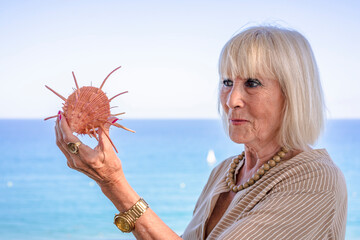 Senior woman looking at a conch or seashell. The woman is dressed in a beige dress and has straight blonde hair