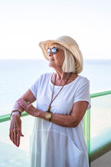 Senior woman leaning on a terrace railing looking out to sea wearing a straw hat and white dress