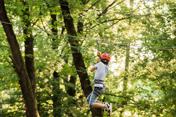 Naklejka na ściany i meble Little boy overcomes the obstacle in the rope park. Climbing in high rope course enjoying the adventure. Adventure climbing high wire forest - people on course in mountain helmet and safety equipment.
