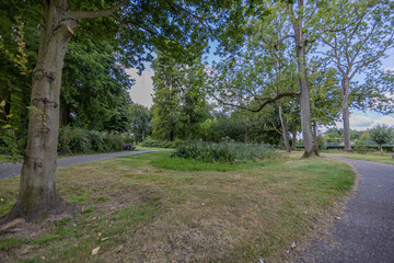 Sittard city park with lush trees, green grass, two pedestrian paths, a wooden bench on a sunny summer day in South Limburg, the Netherlands