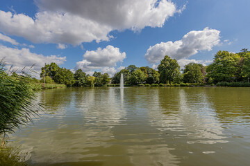 Lake with a fountain with a jet of water surrounded by lush green trees in the city park of Sittard, sunny summer day with a blue sky and white clouds in South Limburg, the Netherlands