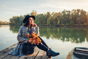 Woman relaxing on lake pier by boat admiring autumn landscape holding branches with leaves. Fall season activities © maryviolet