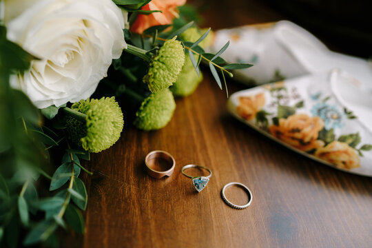 Wedding rings and an engagement ring on a wooden table with women's wedding shoes and a bouquet of flowers.
