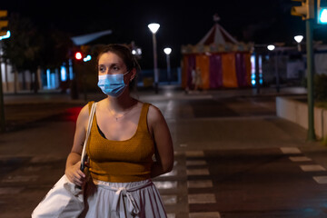 Young Hispanic woman crossing a crosswalk at night alone with a mask against Covid19, wears a brown top with white shorts and carries a cloth bag