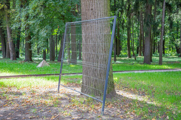 Part of a fence made of iron gray mesh and a tree trunk in a city park