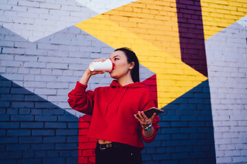 Good looking female millennial drinking tasty coffee from takeaway cup, Chinese generation Y with cellphone technology in hand enjoying caffeine beverage at urbanity with colorful street art © BullRun