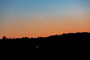 A young moon on an orange-blue sunset sky over a dark forest. Horizontal orientation. High quality photo