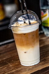 Ice latte coffee drink in a transparent plastic disposable cup with two black plastic tubes on a wooden counter outside. Vertical orientation. High quality photo