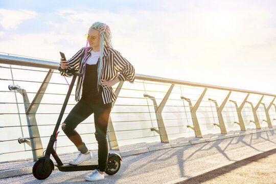 Girl with colored dreadlocks is standing on bridge, holding smartphone. Young woman in yellow glasses came to meeting by electric scooter. Moving by ecological eco portable transport of future concept