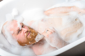 Close-up portrait of funny red-bearded bald man taking a bath with foam. Cheerful naughty guy at...