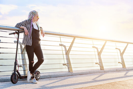 Girl with colored dreadlocks is standing on bridge in modern city. Young woman in yellow glasses came to meeting by electric scooter. Moving by ecological eco portable transport of future concept.