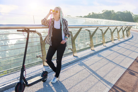 Girl with colored dreadlocks is standing on bridge in modern city. Young woman in yellow glasses came to meeting by electric scooter. Moving by ecological eco portable transport of future concept.