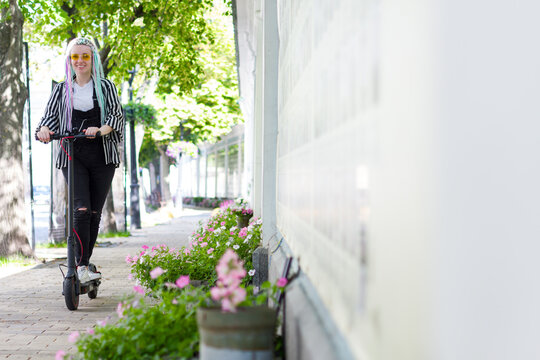 Smiling girl with colorful dreadlocks is riding, driving electric scooter in street of city. Young woman is walking in old town. Moving by ecological eco portable transport of future concept.