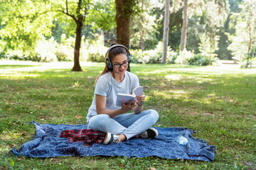 Obraz premium Young woman sitting in the park reading the book and listening to music over the wireless headphones