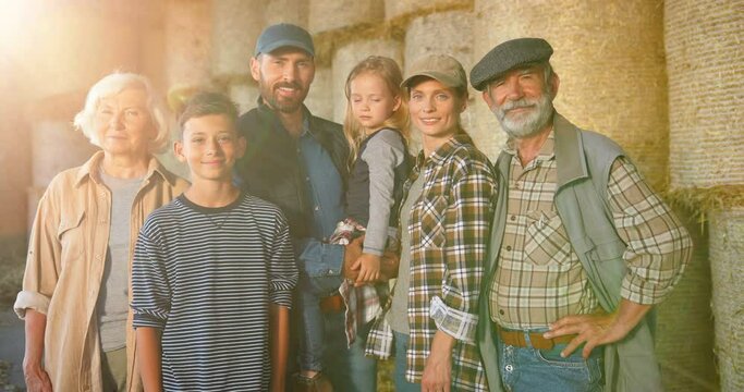 Portrait of happy Caucasian family of three generations standing outdoor at hay stocks and smiling to camera. Old parents with children and grandchildren in countryside. Farmers at farm. Summer.