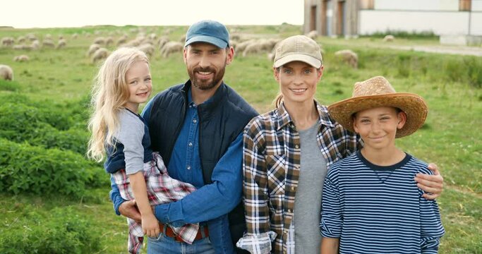 Portrait of Caucasian happy family with little kids standing at pasture with sheep flock on background and smiling to camera. Cheerful parents with small son and daughter in field at farm.
