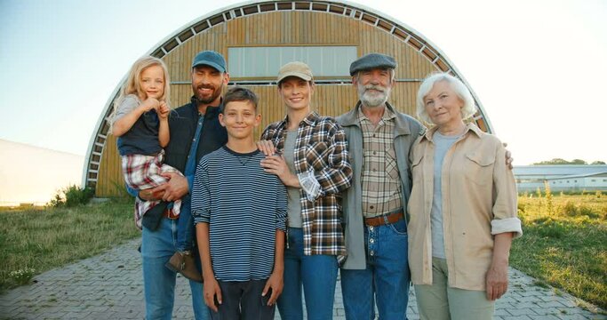Portrait happy Caucasian family of three generations standing outdoor at farm shed and smiling to camera. Old parents with children and grandchildren in countryside. Farmers. Summer. Village lifestyle