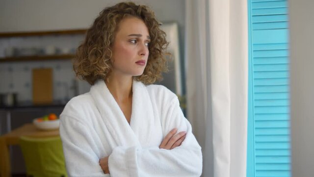 Sad young woman in bathrobe standing at window and looking into distance