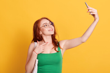 Portrait of young attractive woman making selfie photo with smart phone isolated over yellow background, red haired female in green t shirt pointing to phone's camera with index finger.