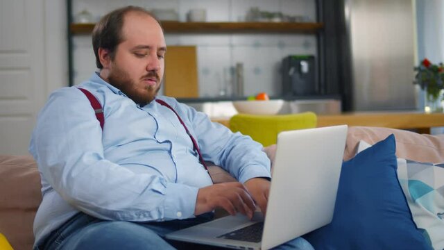 Funny fat man working on laptop sitting on couch at home