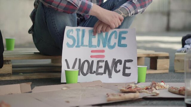 Close-up of Silence is violence banner with unrecognizable male protester sitting on pallet at the background. Caucasian man with placard against gender or ethnic inequality.