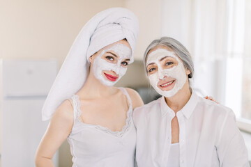 Portrait of two Caucasian women, young girl with hair wrapped in towel, and elderly gray haired woman, with homemade facial masks on faces, smiling to camera, posing at home. Beauty treatment