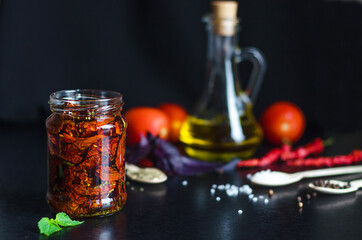 Homemade sun-dried red tomato slices in glass jar with olive oil, basil, oregano spices on dark background. Cooking process traditional Italian Mediterranean cuisine. copy space, text